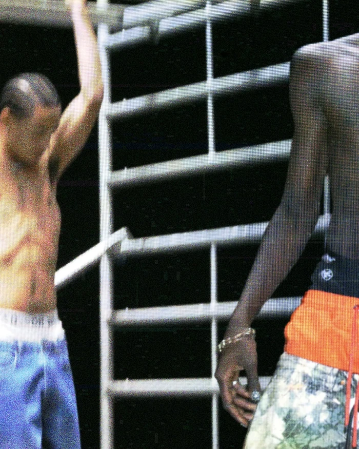 Two shirtless young men wearing low-rise streetwear pants and shorts posing near white metal scaffolding against a dark backdrop.
