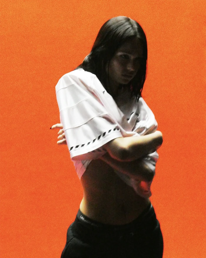 A model lifts a white graphic jersey to expose their midriff against a vibrant solid orange studio backdrop.