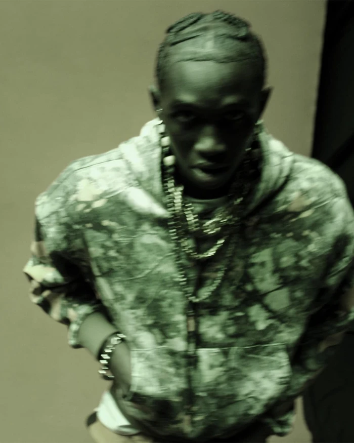 Young Black man in a camo zip hoodie with layered chain necklaces looking up at the camera against a plain backdrop.