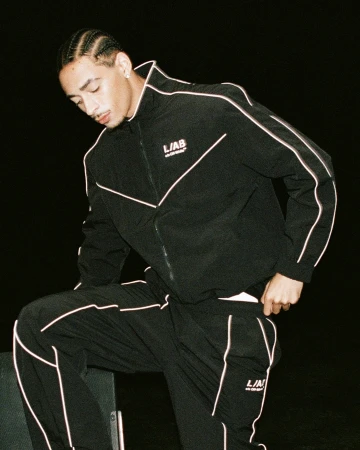 A young man with braided hair wearing a black Off-White tracksuit with light piping against a dark backdrop.