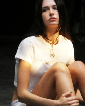 Woman wearing a white cropped t-shirt and metallic necklace seated against a dark studio background.