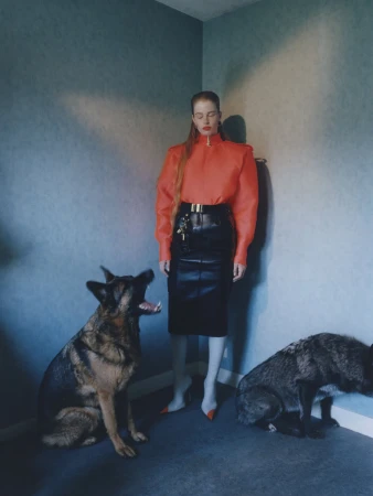 Vivian Jenna Wilson poses in an orange high-neck top and black leather skirt in a corner flanked by two German Shepherd dogs.