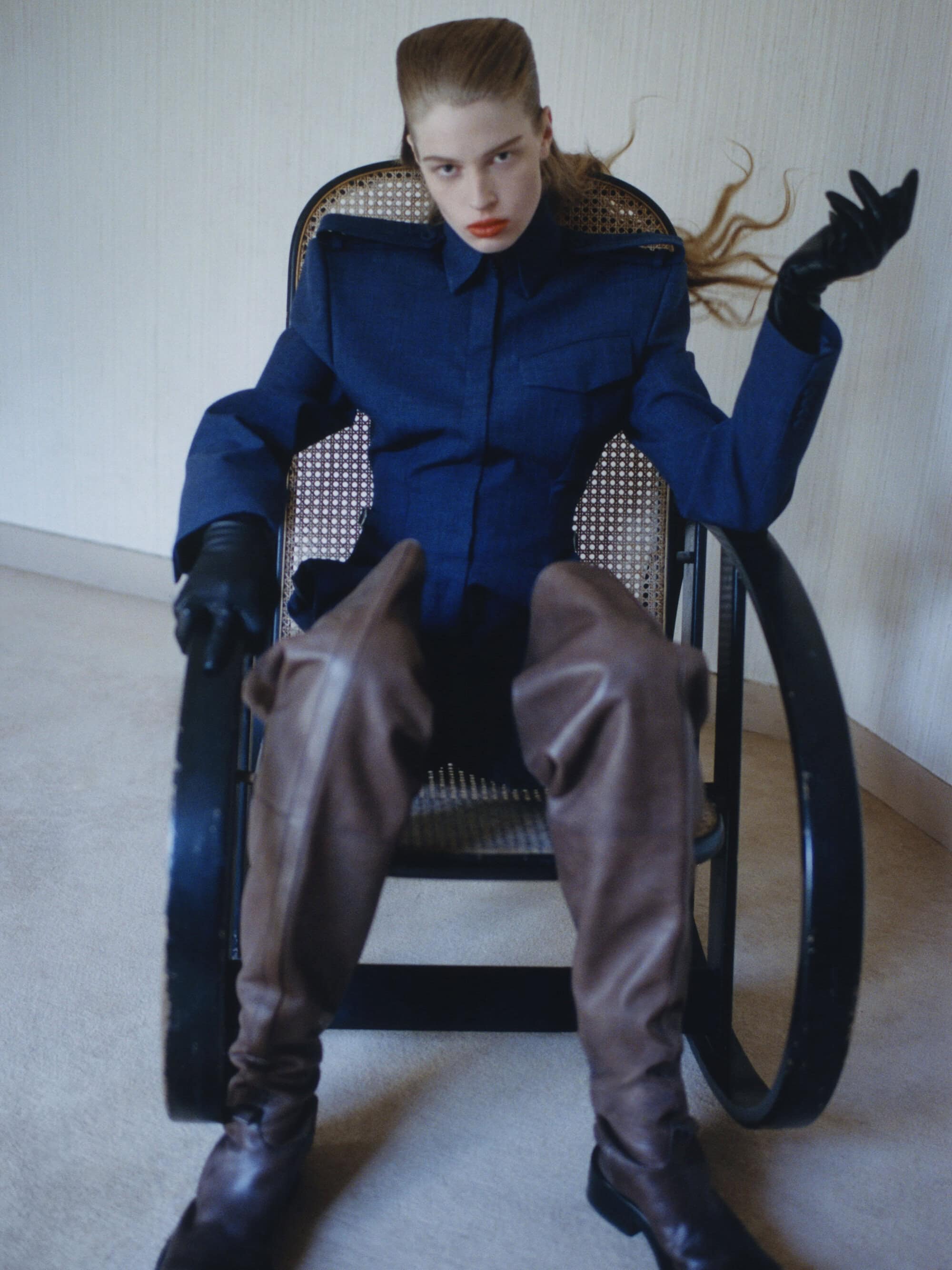 Vivian Jenna Wilson in a structured navy jacket and brown leather thigh-high boots seated in a black cane rocking chair.