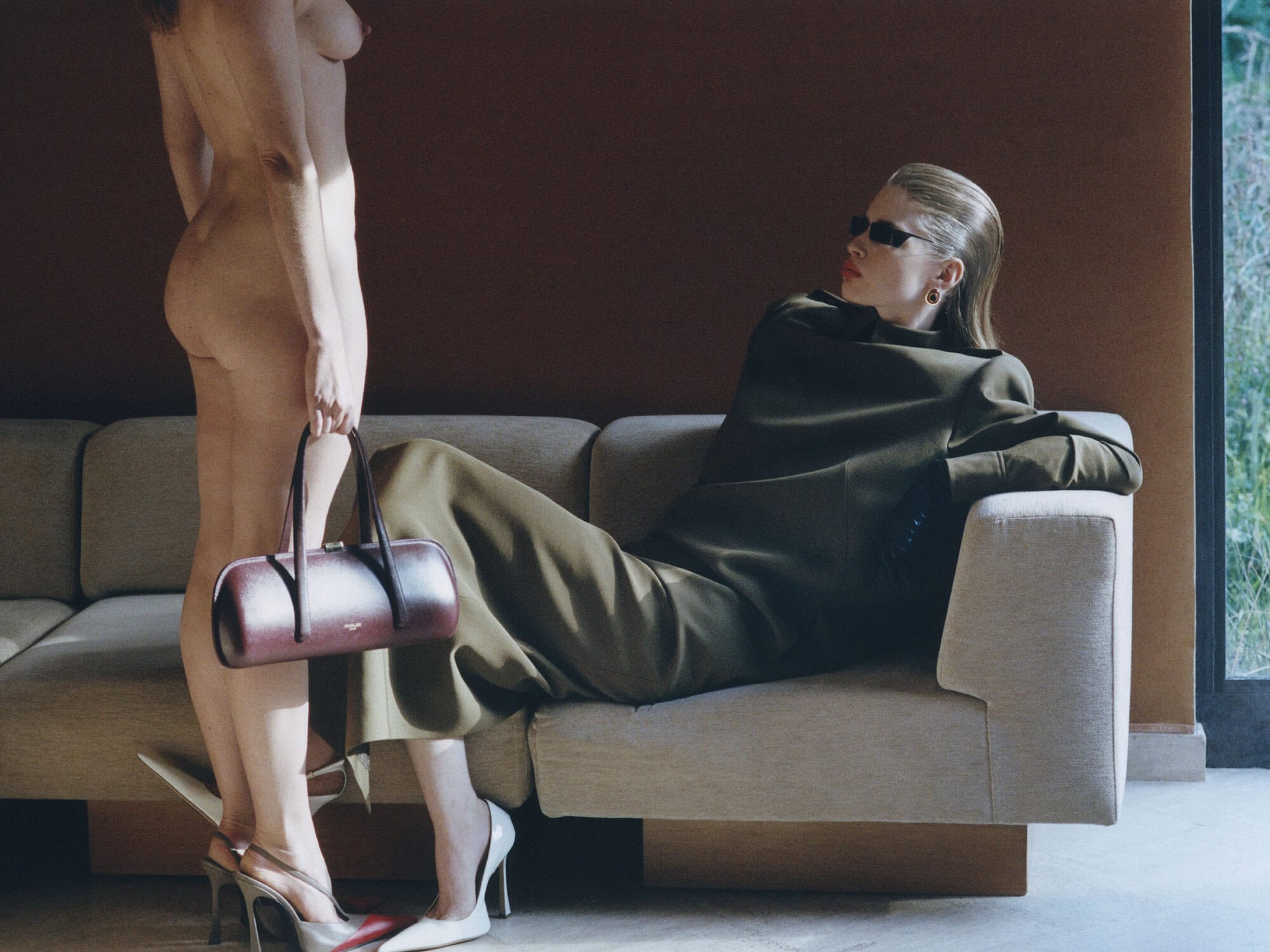 Model reclining on a beige sofa in an olive green coat and sunglasses beside a standing nude figure holding a leather handbag.