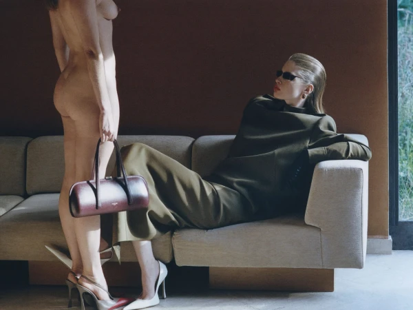 Model reclining on a beige sofa in an olive green coat and sunglasses beside a standing nude figure holding a leather handbag.