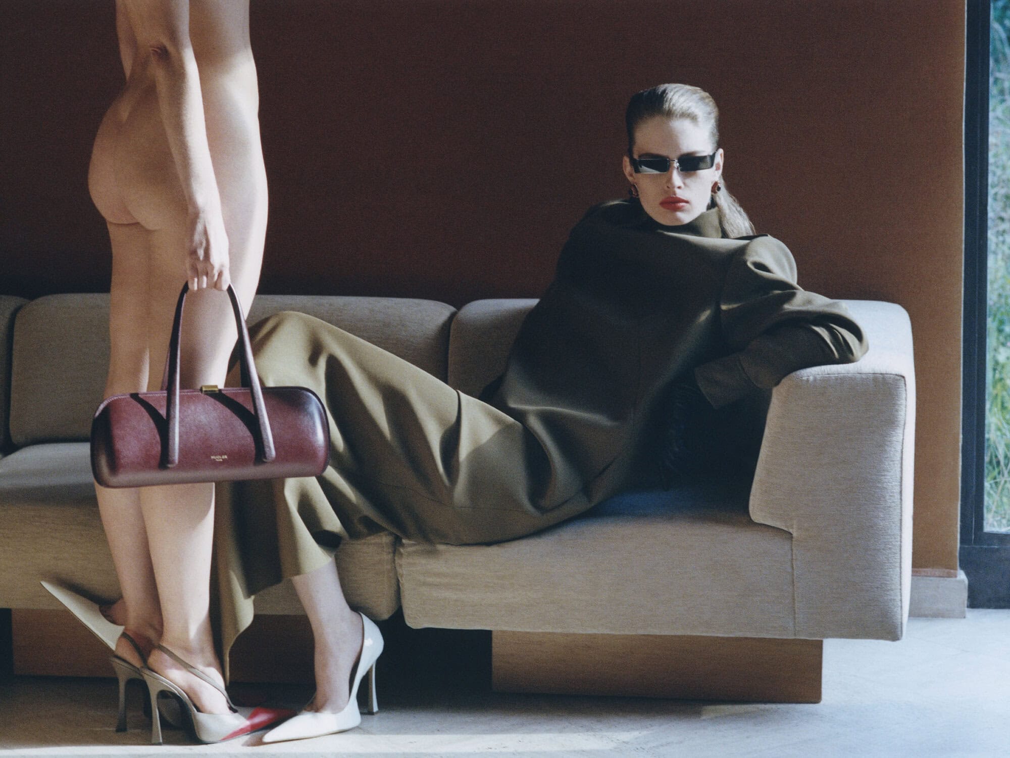 Model Vivian Jenna Wilson reclining on a beige sofa in an olive coat and sunglasses beside a figure holding a burgundy handbag.