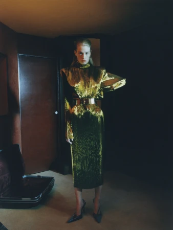 Vivian Jenna Wilson stands in a shimmering metallic gold Mugler dress with structured shoulders in a moody interior.