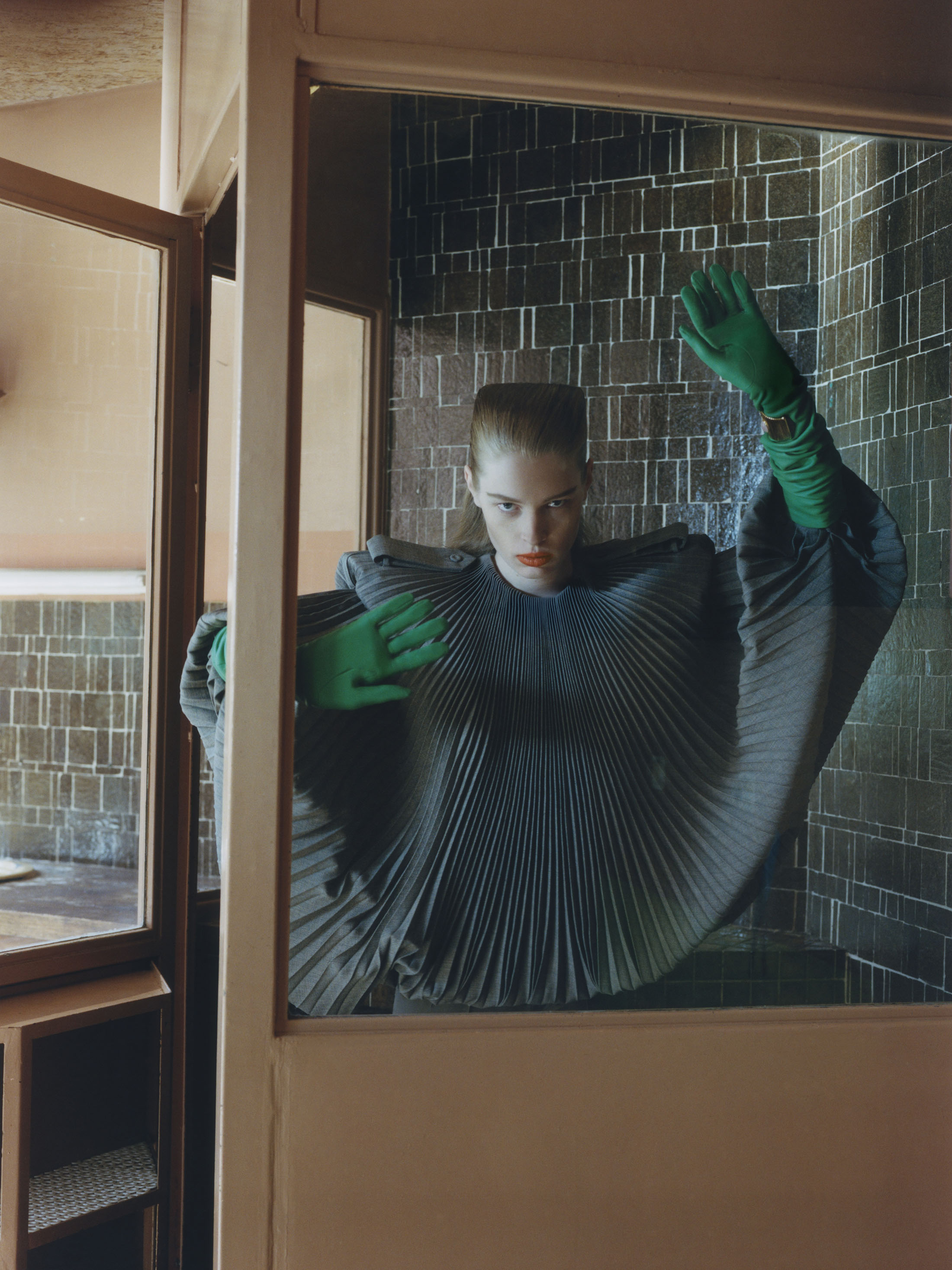 Vivian Jenna Wilson wearing a fan-pleated grey cape and bright green gloves with her hands pressed against a framed glass window pane.