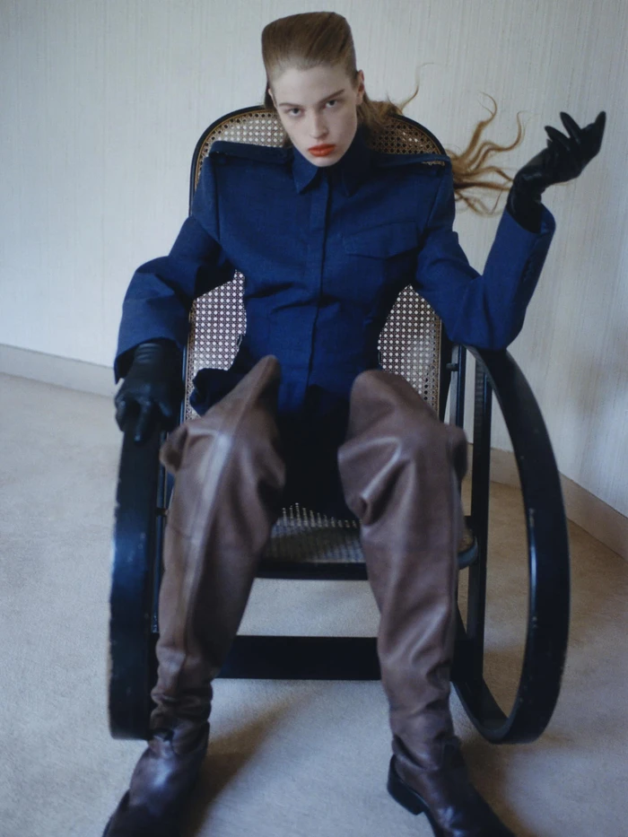 Vivian Jenna Wilson in a structured navy jacket and brown leather thigh-high boots seated in a black cane rocking chair.