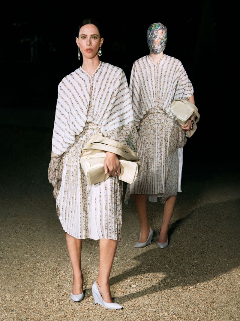 Rebecca Hall standing outdoors at night in a pleated striped cape dress with oversized clutch beside a masked model.