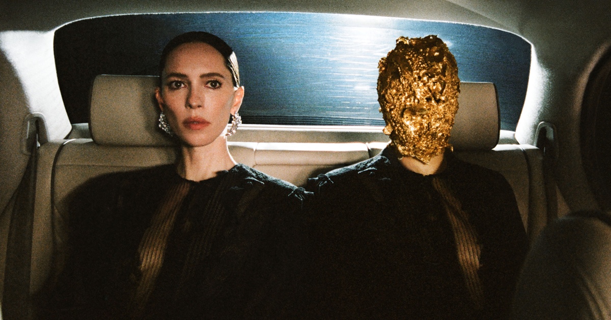 Rebecca Hall seated beside a person wearing a full gold textured mask in the backseat of a car.