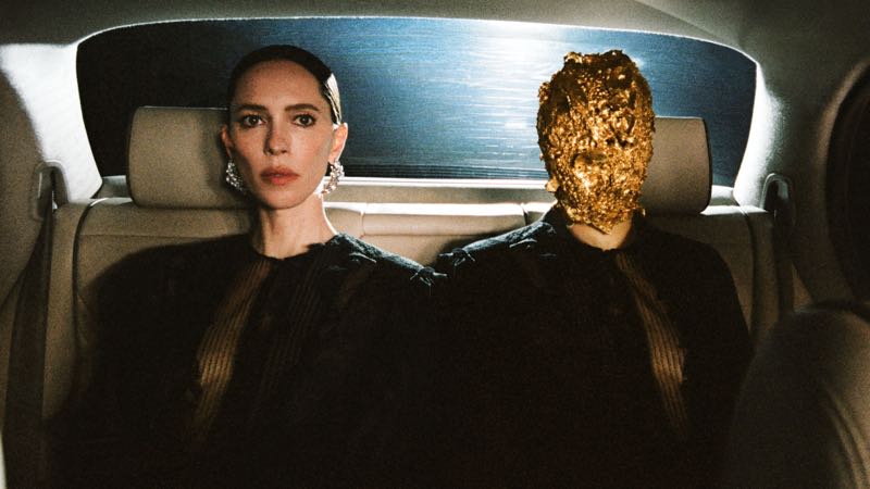 Rebecca Hall seated next to a figure in a gold textured mask in the backseat of a car for Maison Margiela.