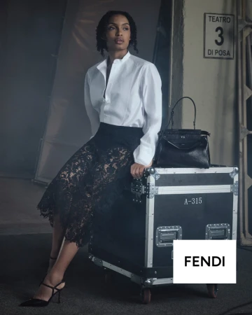 Yara Shahidi seated on a black flight case wearing a white shirt and black lace skirt beside a black Fendi Peekaboo handbag.