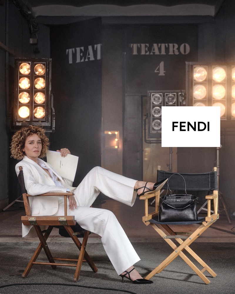 Valeria Golino in a white pantsuit seated across wooden director chairs with a black Fendi Peekaboo handbag on a soundstage set.