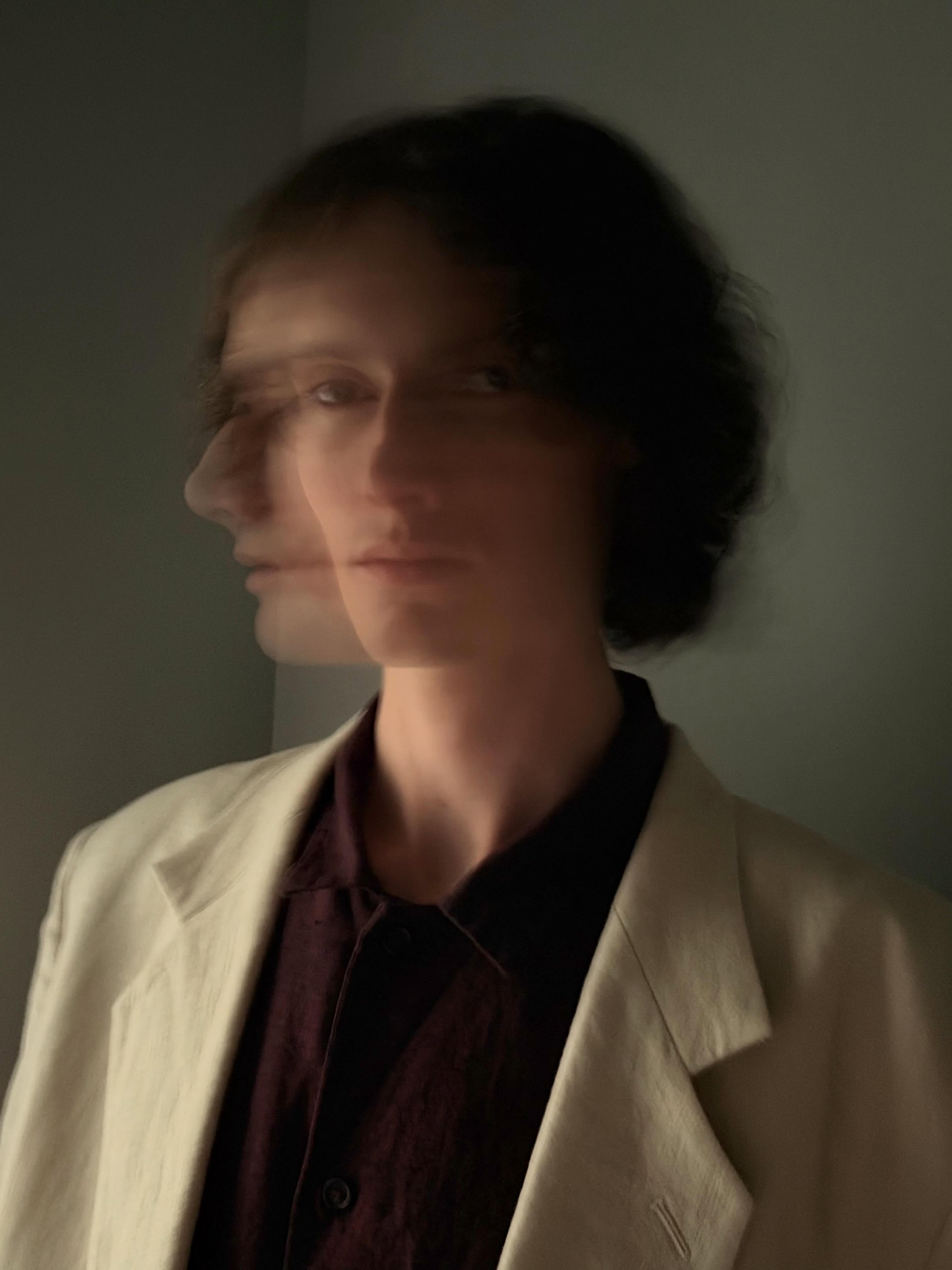 Portrait of a person with motion blur showing double facial exposure, wearing a cream blazer and dark shirt against a dark background.