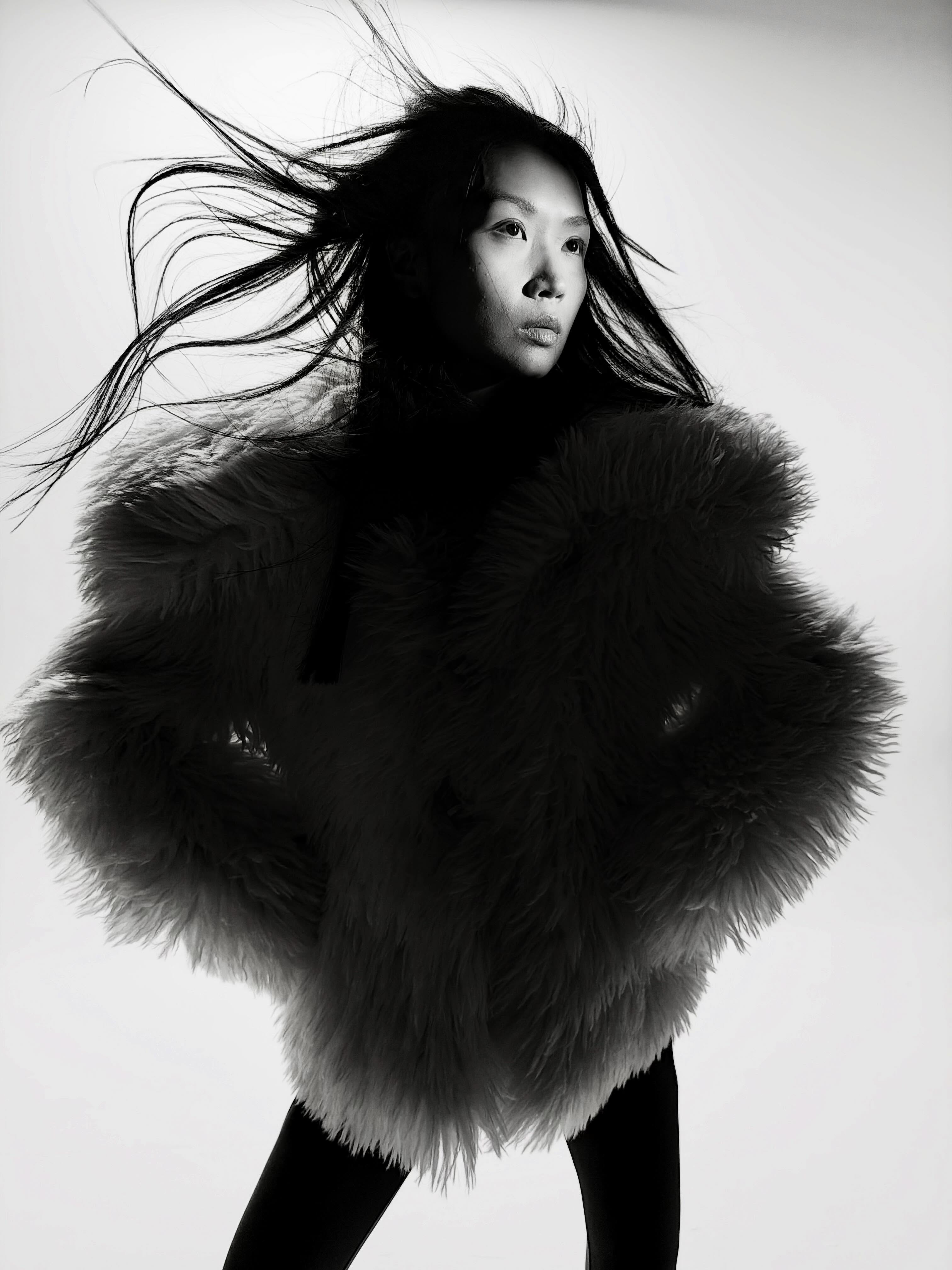 East Asian woman in a voluminous shaggy jacket with windblown hair posed against a white studio background.