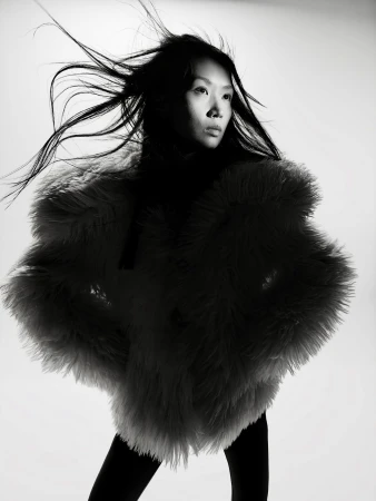 East Asian woman in a voluminous shaggy jacket with windblown hair posed against a white studio background.