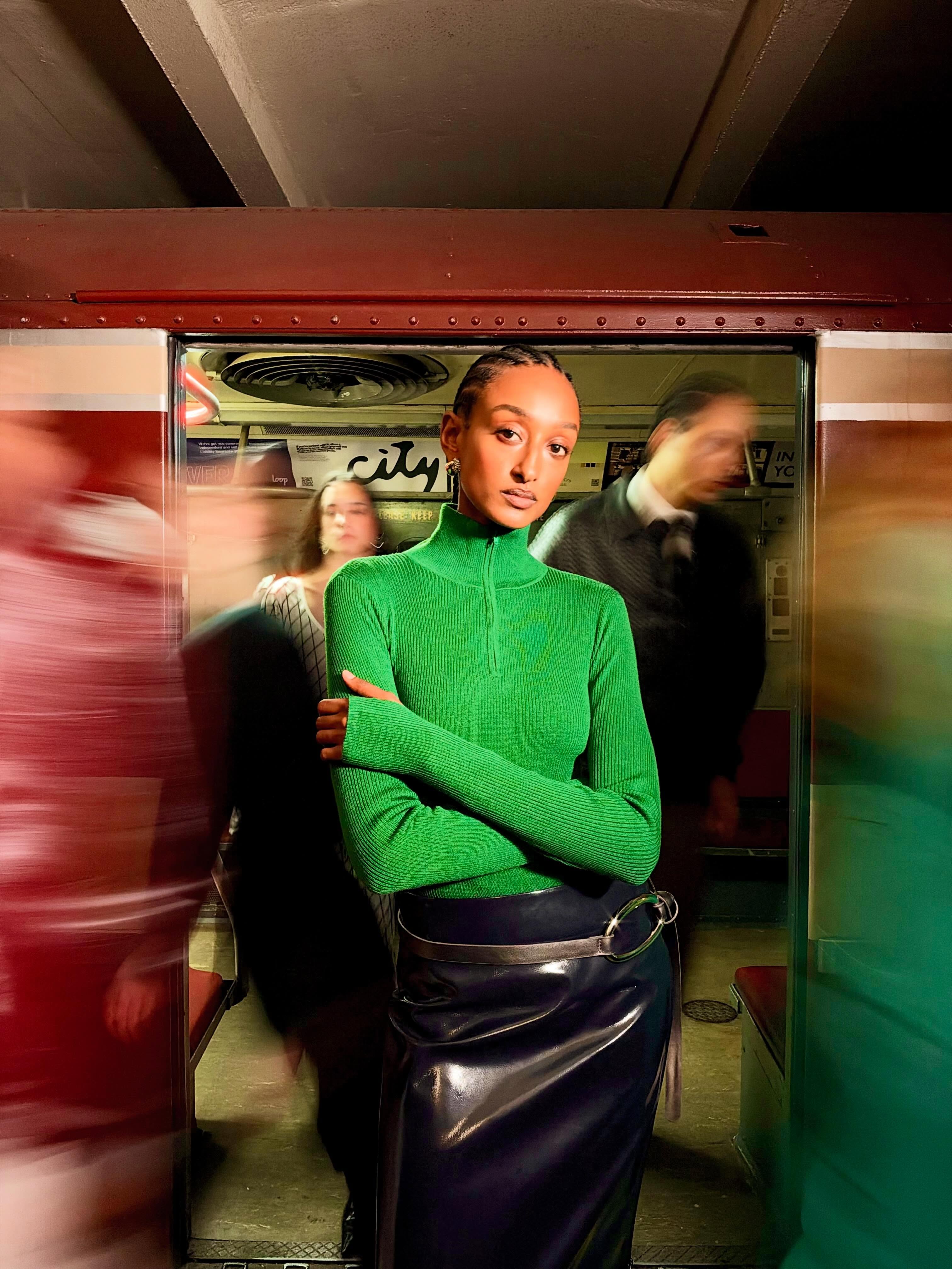 Woman in vibrant green ribbed top and dark glossy skirt stands in a subway doorway surrounded by motion-blurred passengers.