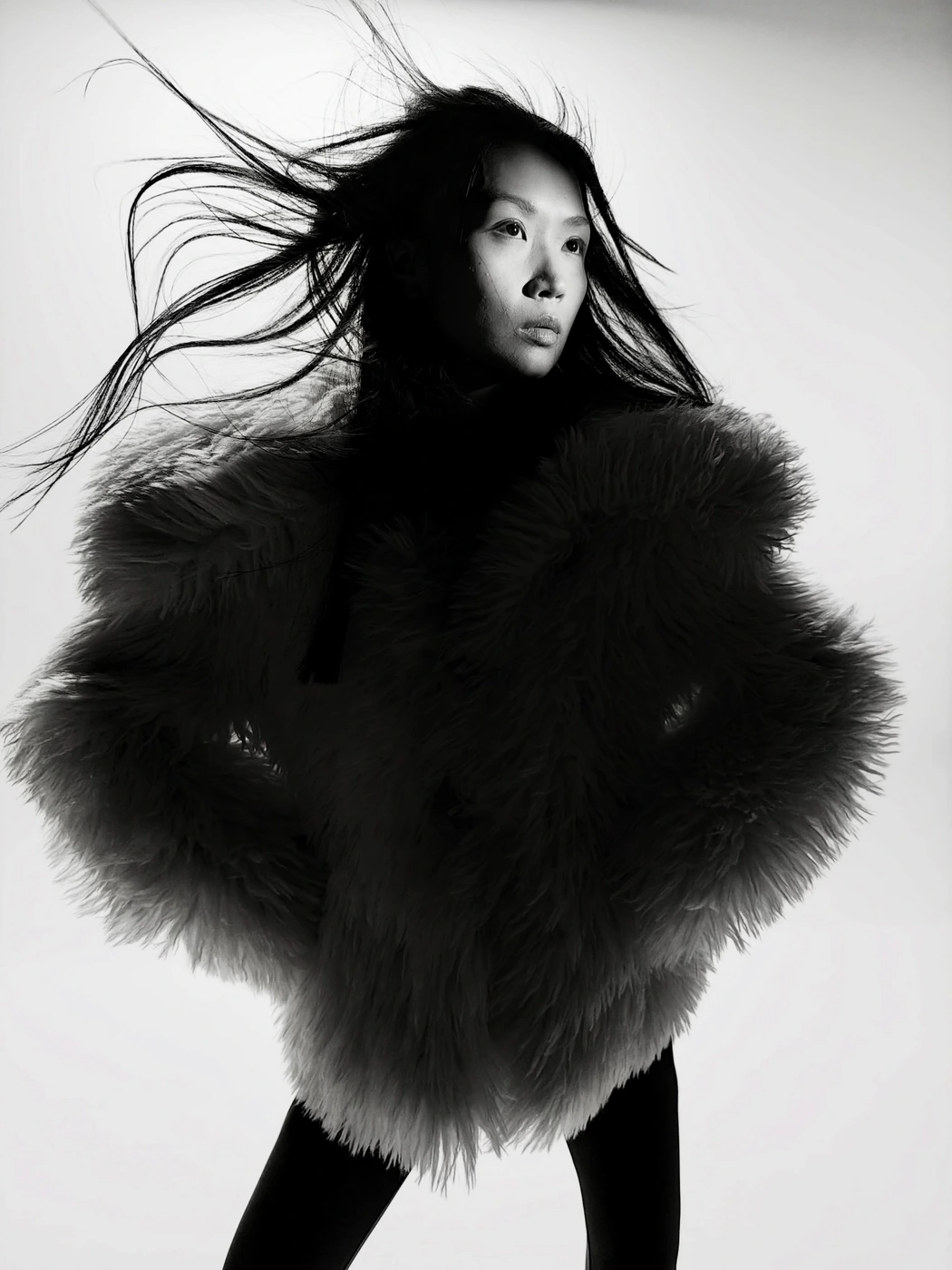 East Asian woman in a voluminous shaggy jacket with windblown hair posed against a white studio background.