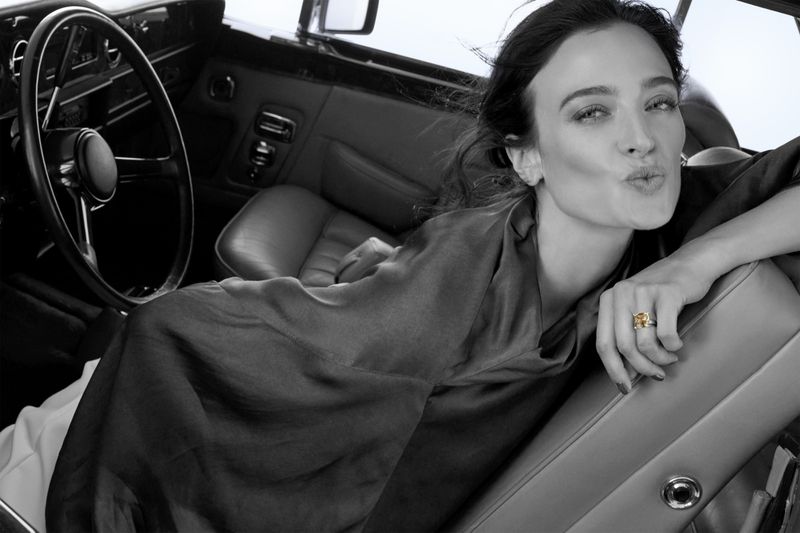 Monochrome portrait of a woman with wavy hair wearing a draped top and a gold ring inside the luxurious leather interior of a car.