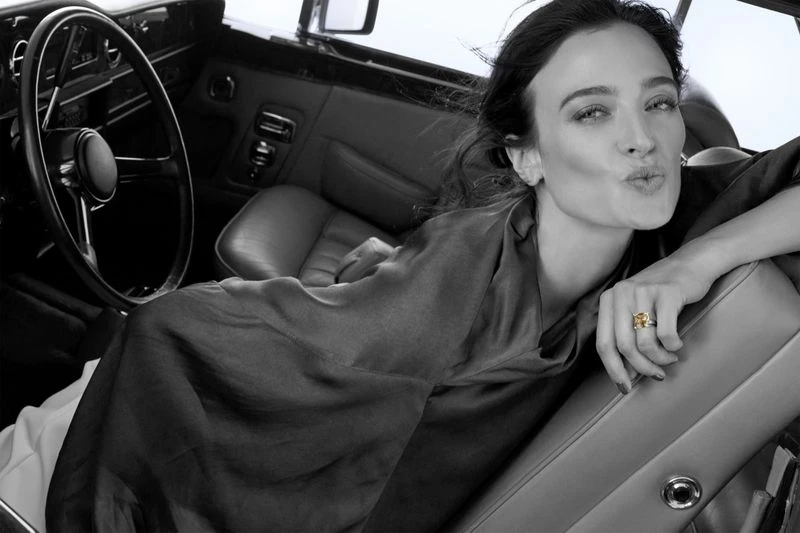 Monochrome portrait of a woman with wavy hair wearing a draped top and a gold ring inside the luxurious leather interior of a car.