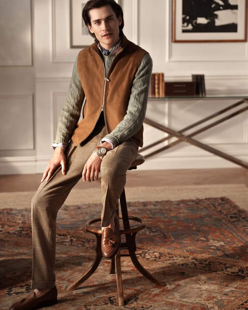 Man in vest and brown pants sitting on wooden stool in interior setting with rug, books, and framed art, styled professionally.