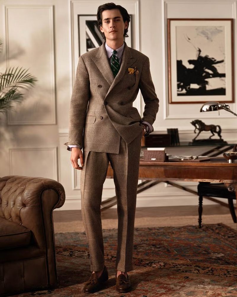 Man in a brown tweed double-breasted suit, green tie, and brown loafers stands in a room with a wooden desk and traditional art.