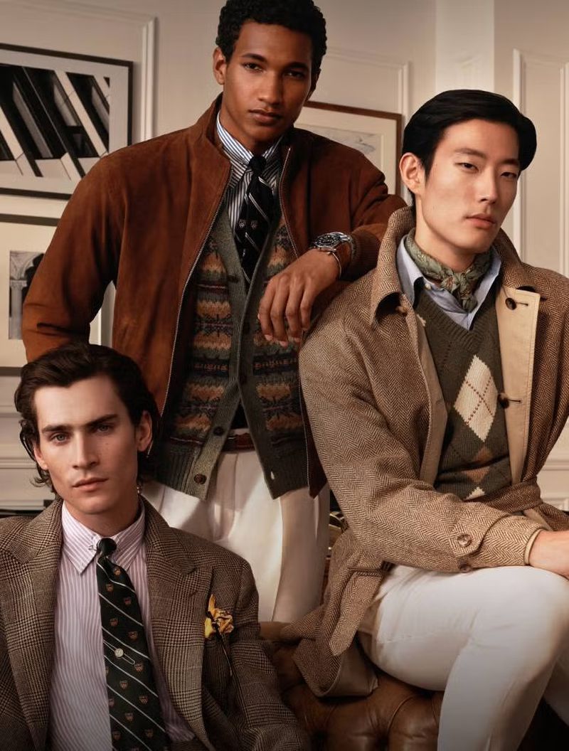 Three men in jackets and ties pose indoors. The man in the center wears a brown bomber jacket and patterned cardigan over a button-down shirt.