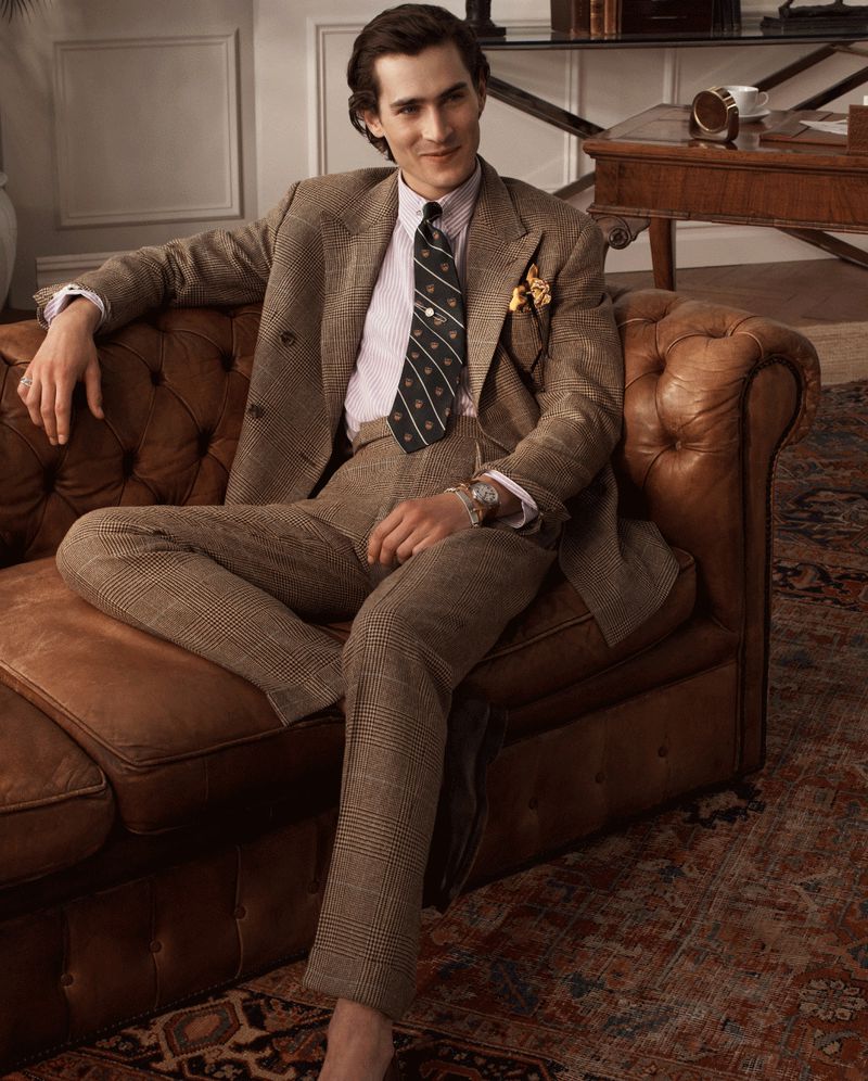 A man in a brown suit sits on a brown leather couch, wearing a tie and dress shoes, and a patterned rug is on the floor.