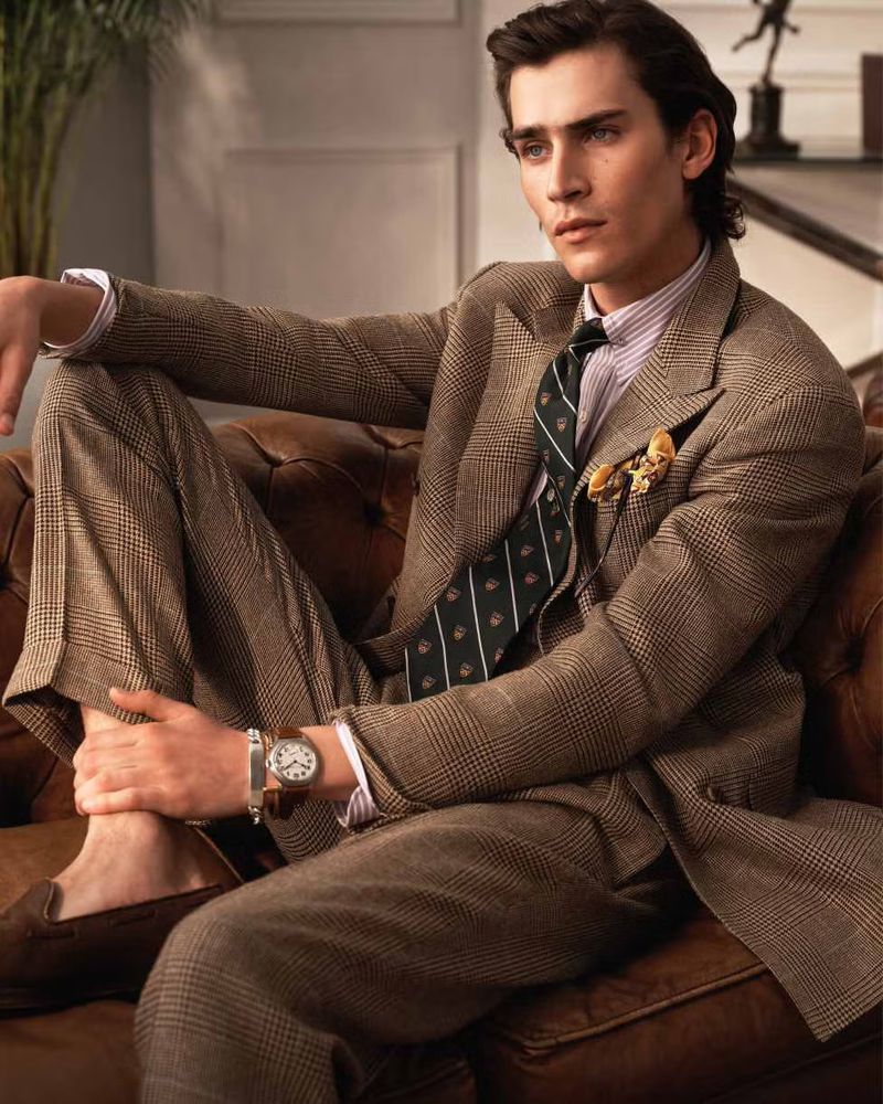 Adult male model lounges on a leather couch wearing a brown patterned suit and tie with moccasins in a casual indoor fashion shot.