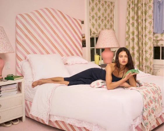 Woman in a navy dress reclining on a pink striped bed holding a small green lizard in a styled bedroom.