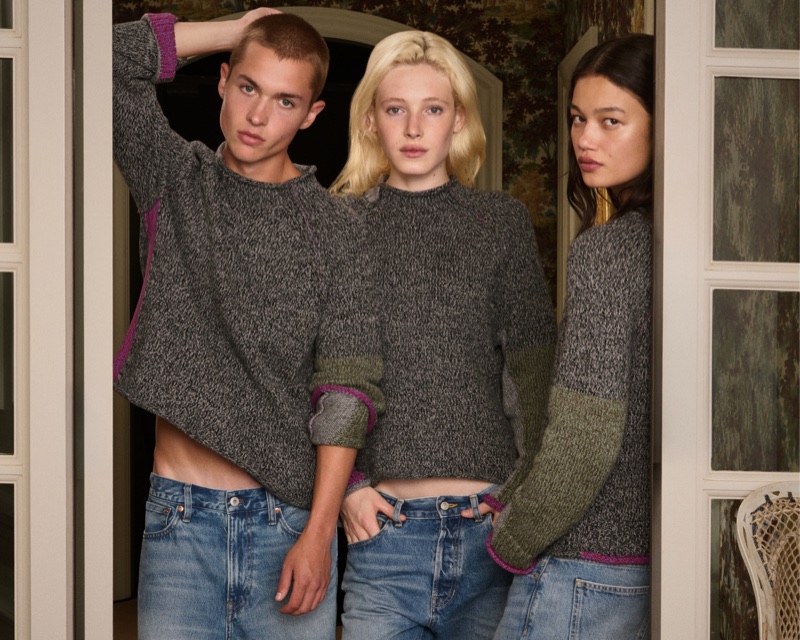 Three models wearing matching grey knit sweaters and blue jeans posing together in a doorway for J.Crew and Eckhaus Latta