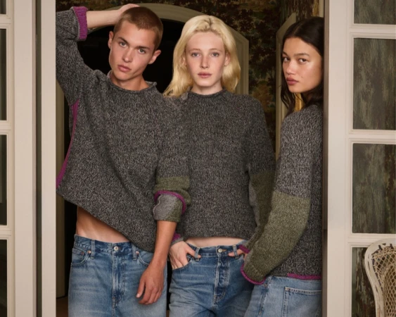 Three models wearing matching grey knit sweaters and blue jeans posing together in a doorway for J.Crew and Eckhaus Latta