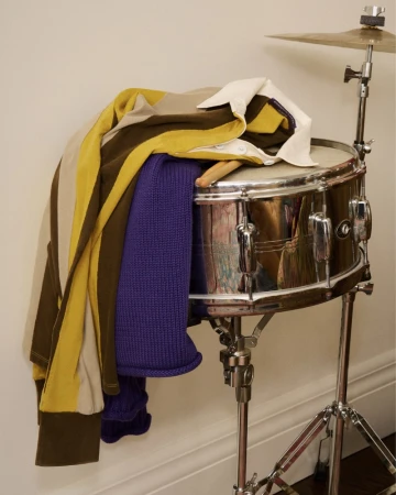 Folded polo shirt and purple knit sweater resting on a chrome snare drum with drumsticks.