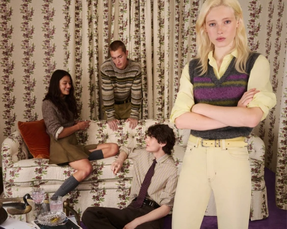 Group of four models posing in a room with patterned floral curtains and sofa, featuring a woman in a yellow sweater vest and pants foreground.