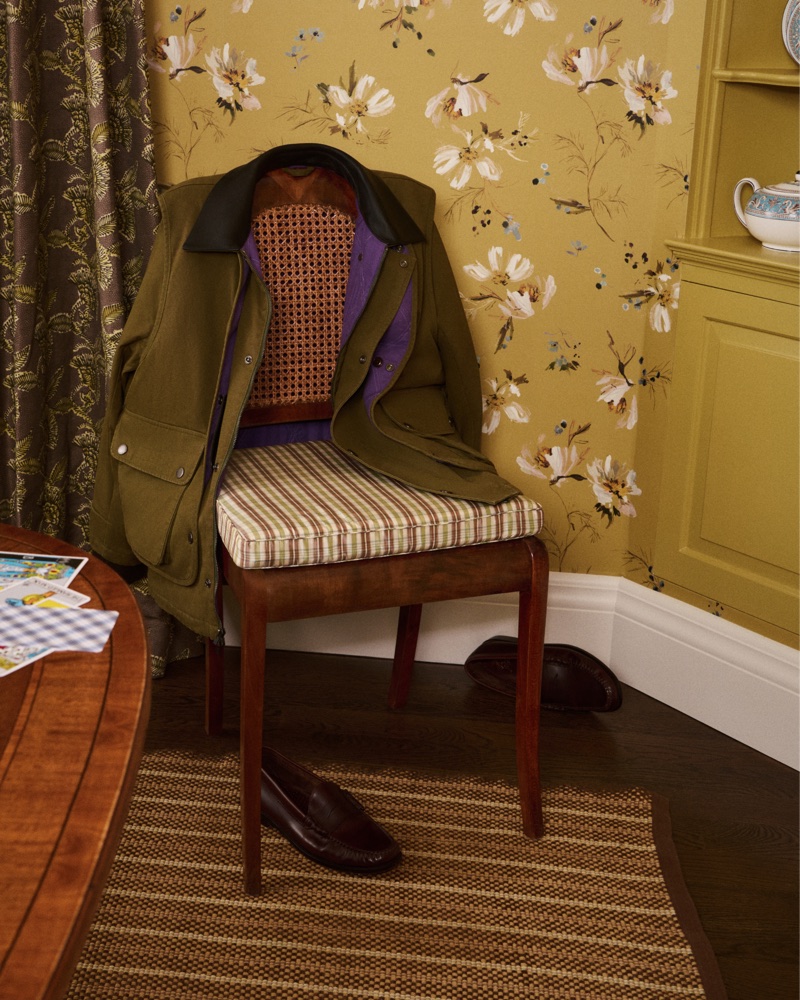 An olive green jacket draped over a wooden chair in a room with floral wallpaper and striped cushion.