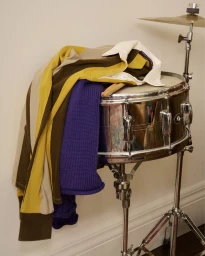 Folded polo shirt and purple knit sweater resting on a chrome snare drum with drumsticks.