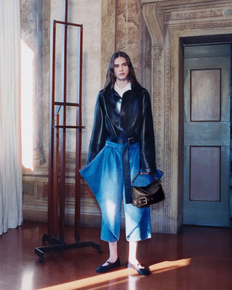 Fashion shot of a young woman indoors wearing a black leather jacket, culottes, and black flats, holding a purse, standing near an easel.