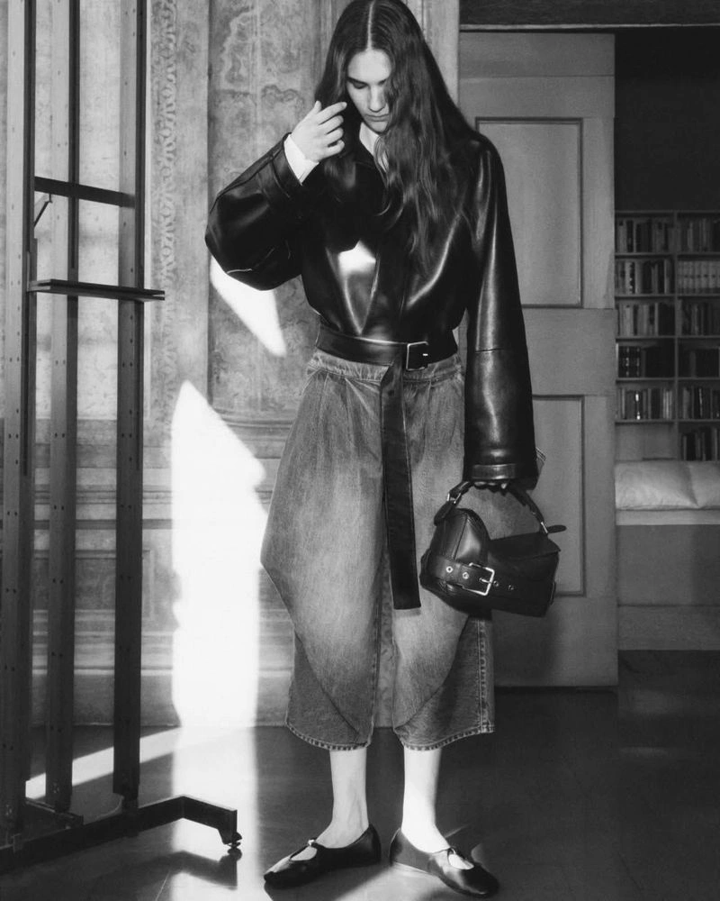 Model in black leather jacket and wide-leg trousers holding a handbag in a room