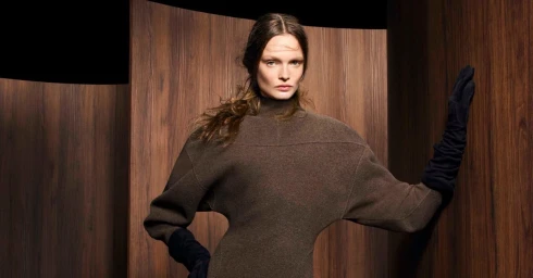 Model Lulu Tenney wearing a brown high-neck knit dress with long black gloves, posing against curved dark wood paneling.