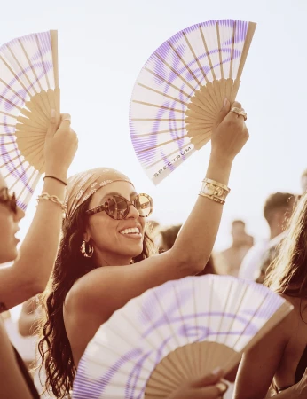 Smiling woman wearing patterned headscarf and round sunglasses holding a folding hand fan in a crowd