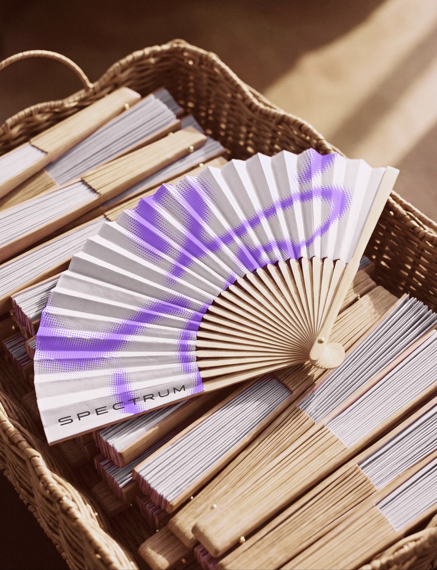 An open wooden folding fan printed with purple graphics and the word SPECTRUM resting atop folded fans in a woven wicker basket.