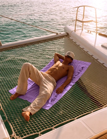 Shirtless man lying on a purple beach towel spread across a catamaran trampoline net over turquoise ocean water.