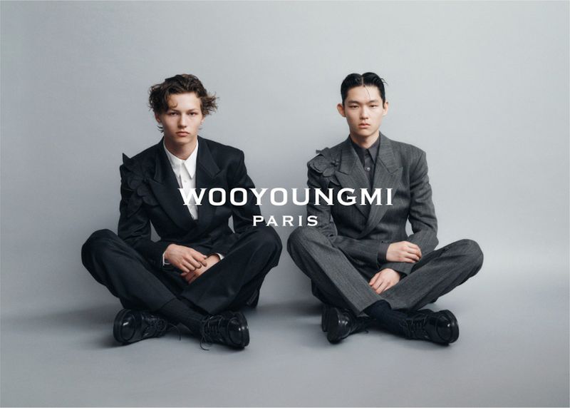 Two fashion models in suits seated on the floor in a photo studio, showcasing designs by Wooyoungmi Paris with studio lighting.