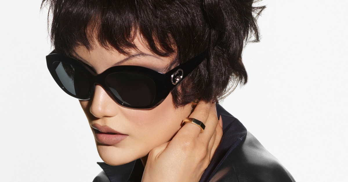 Close-up of a woman with short dark hair wearing oversized black sunglasses and a gold ring against a white background.