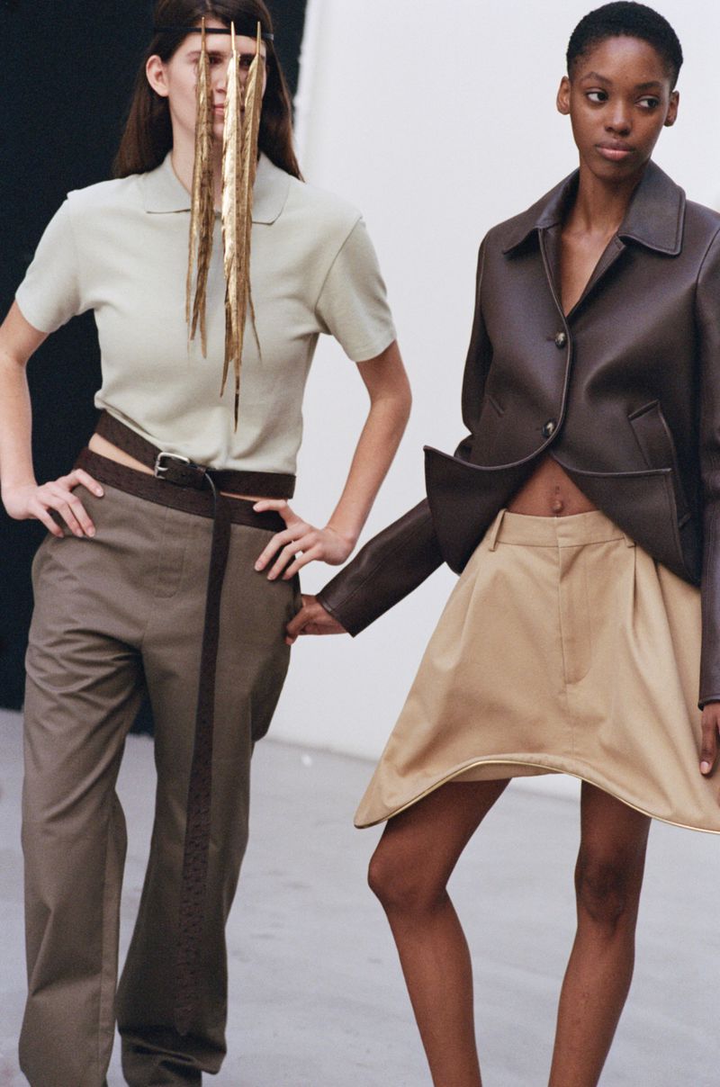 Two fashion models in a studio setting, wearing contemporary apparel, one with a unique fringed headpiece, showcasing minimalist aesthetics.