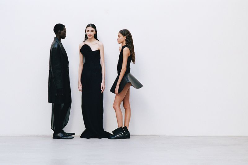 Three fashion models in black dresses and boots pose against a stark white background in a fashion or editorial shoot.