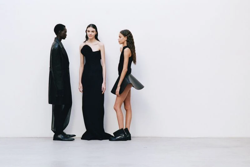 Three fashion models in black dresses and boots pose against a stark white background in a fashion or editorial shoot.