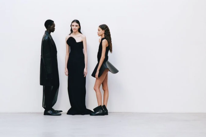 Three fashion models in black dresses and boots pose against a stark white background in a fashion or editorial shoot.