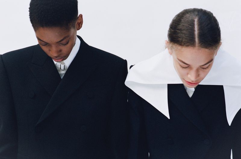Two young fashion models in black blazers, standing side-by-side against a white background, showcasing a minimalist and modern aesthetic.
